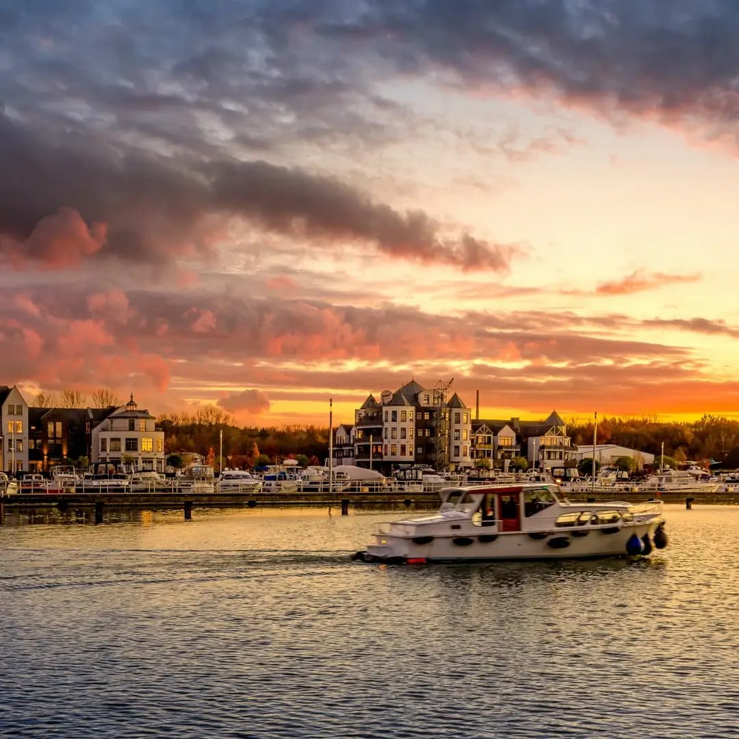 Ein weißes Motorboot fährt bei Sonnenuntergang auf einem Fluss, im Hintergrund ein Hafen mit Gebäuden und orange-grauen Wolken.
