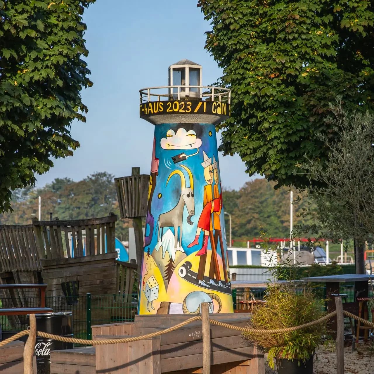 Bunter Leuchtturm mit Graffiti-Kunst, umgeben von Bäumen und Holzkonstruktionen, im Hintergrund ein blauer Himmel.