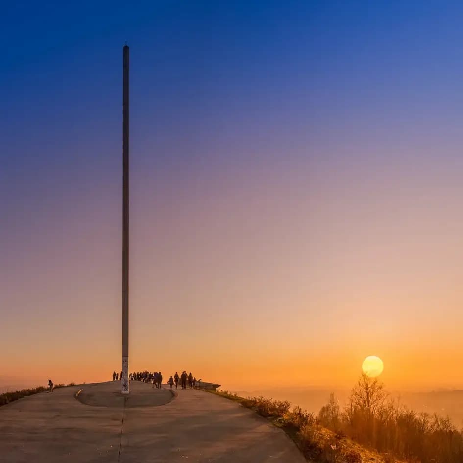 Sonnenuntergang über einer Stadt, mit einem hohen Mast und Menschen auf einem Hügel, die die Aussicht genießen.